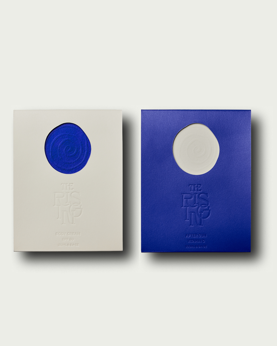 Two pebble-shaped skincare products, one gray and one blue, with 'The Rising' branding on a light gray background.