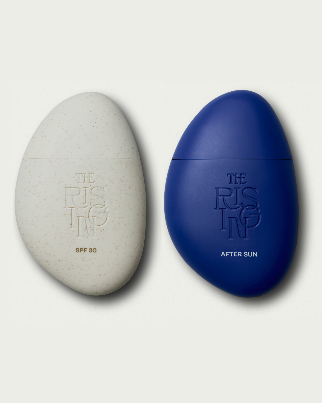 Two pebble-shaped skincare products, one gray and one blue, with 'The Rising' branding on a light gray background.