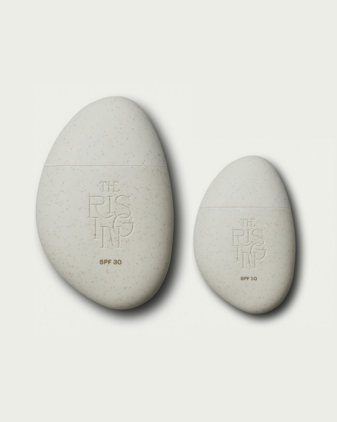 Two pebble-shaped sunscreen products with 'The Rising' branding on a light gray background.