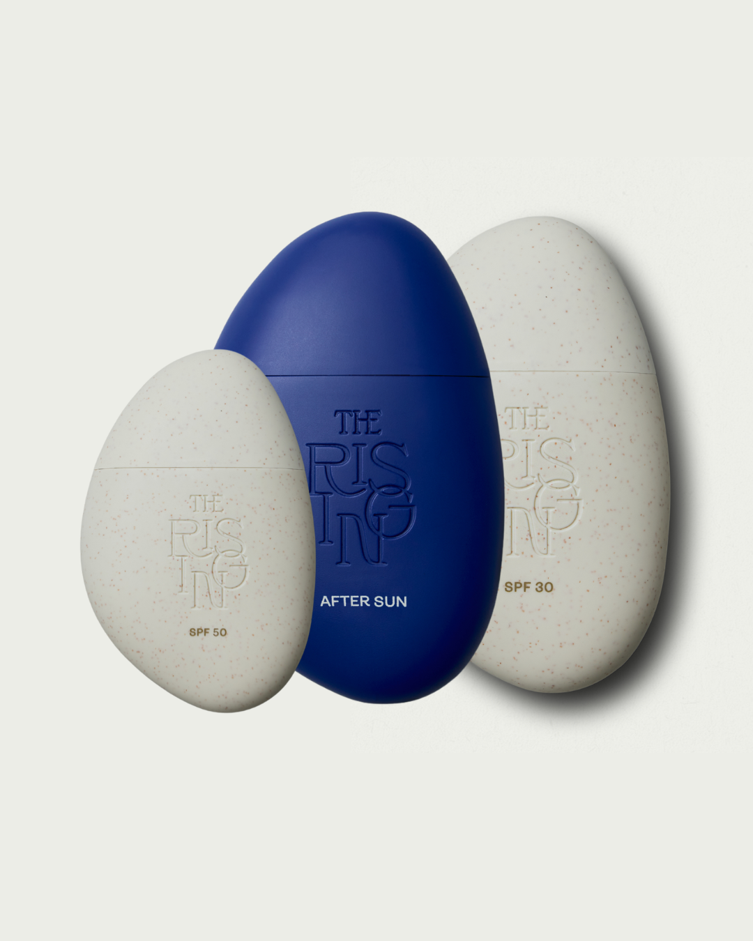 Three pebble-shaped bottles of suncare for face and body and after sun with 'The Rising' branding on a light gray background