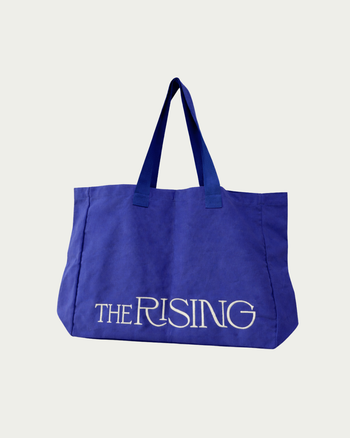 Blue tote bag with 'The Rising' text on a light gray background